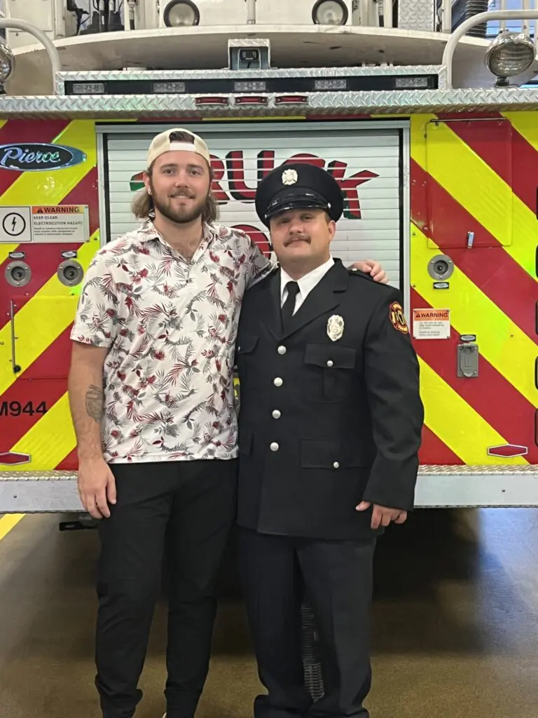 Austin and his brother Paul are standing behind the fire truck at the fire department where he is stationed at 